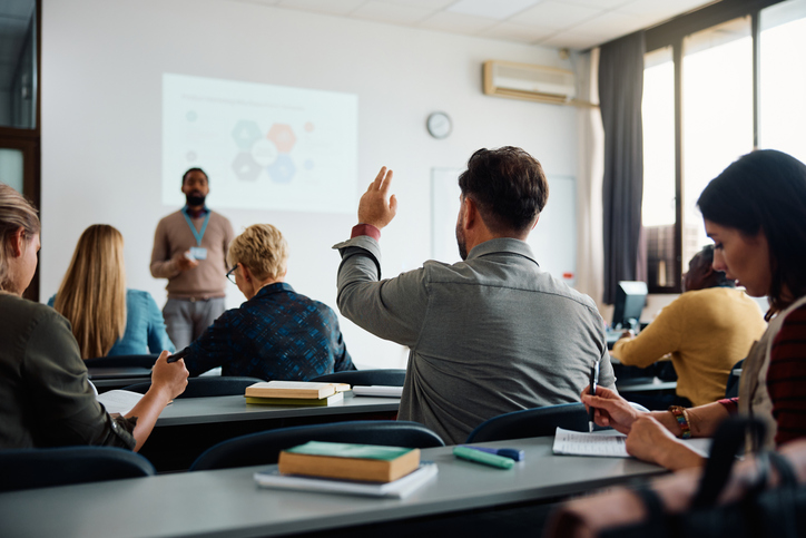 Classroom filled with students talking to a teacher with one student raising his hand. 