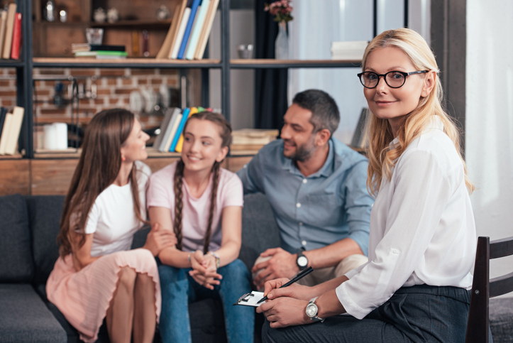 Smiling female therapist looking at camera while happy family with daughter on therapy session image.